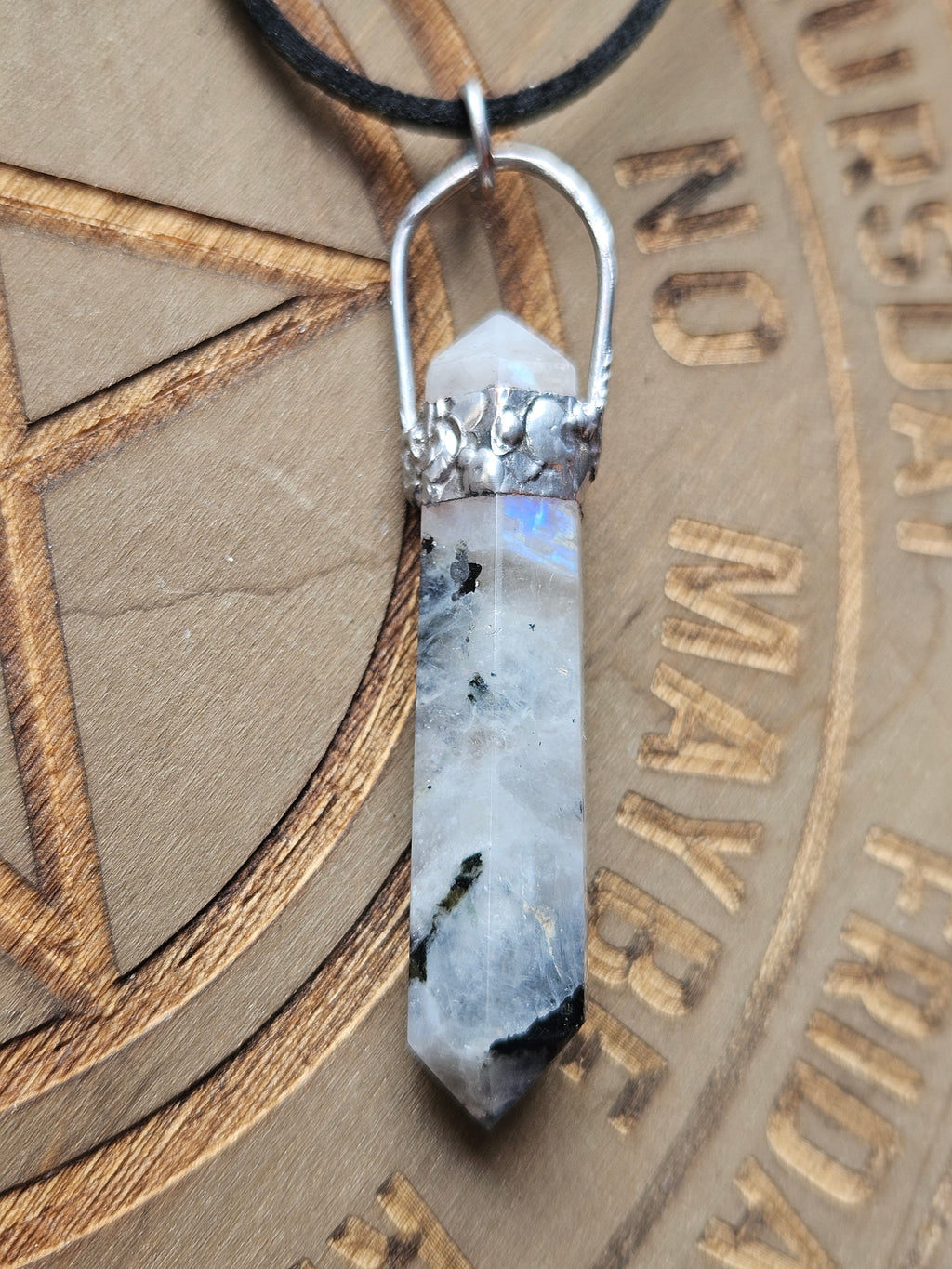 A polished crystal point pendant with a silver metal cap and a black cord, resting on a wooden surface with engraved text.