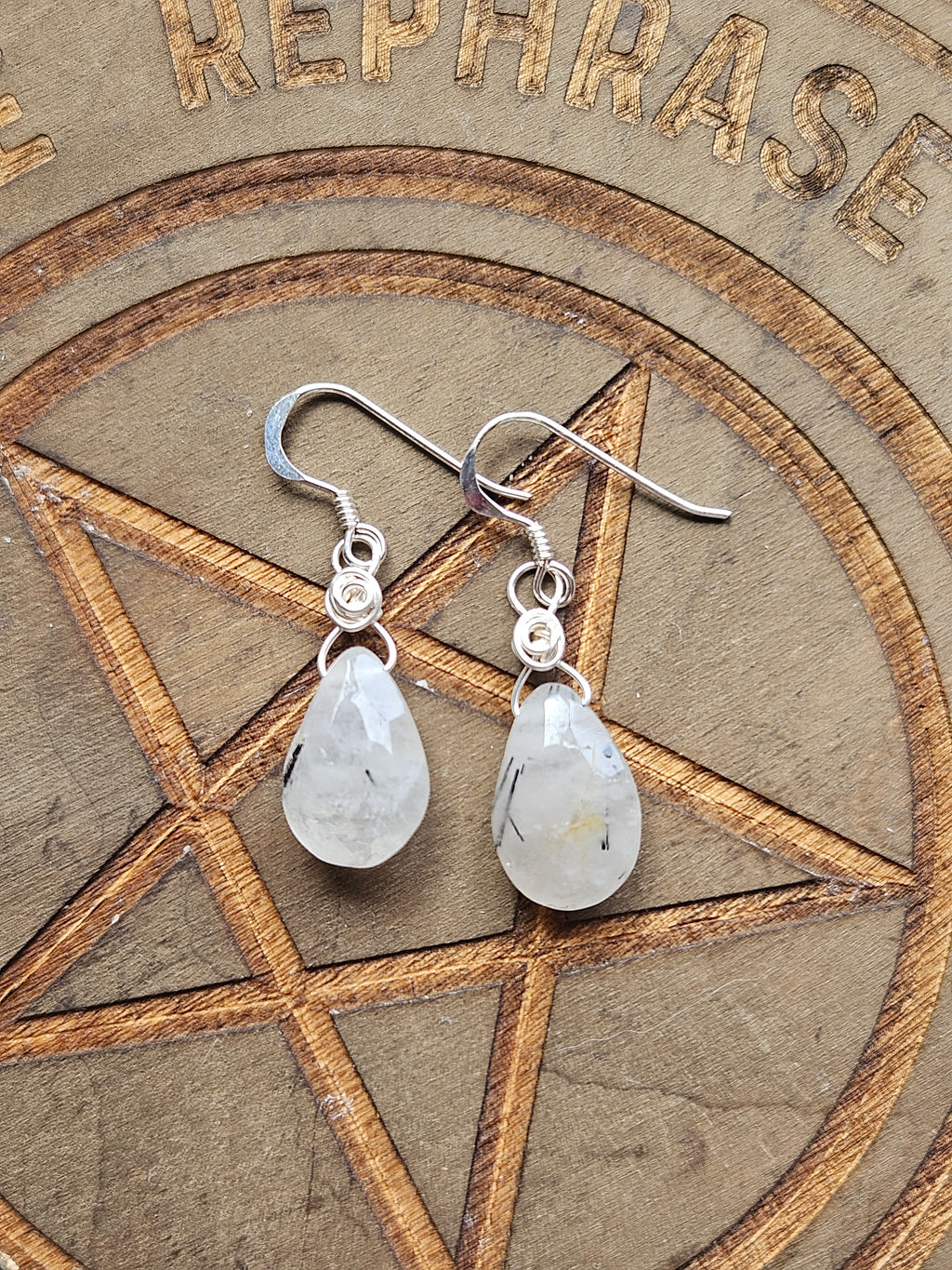 A pair of Sterling Silver dangle earrings with teardrop-shaped Tourmaline In Quartz stones, displayed on a brown circular pentagram background.