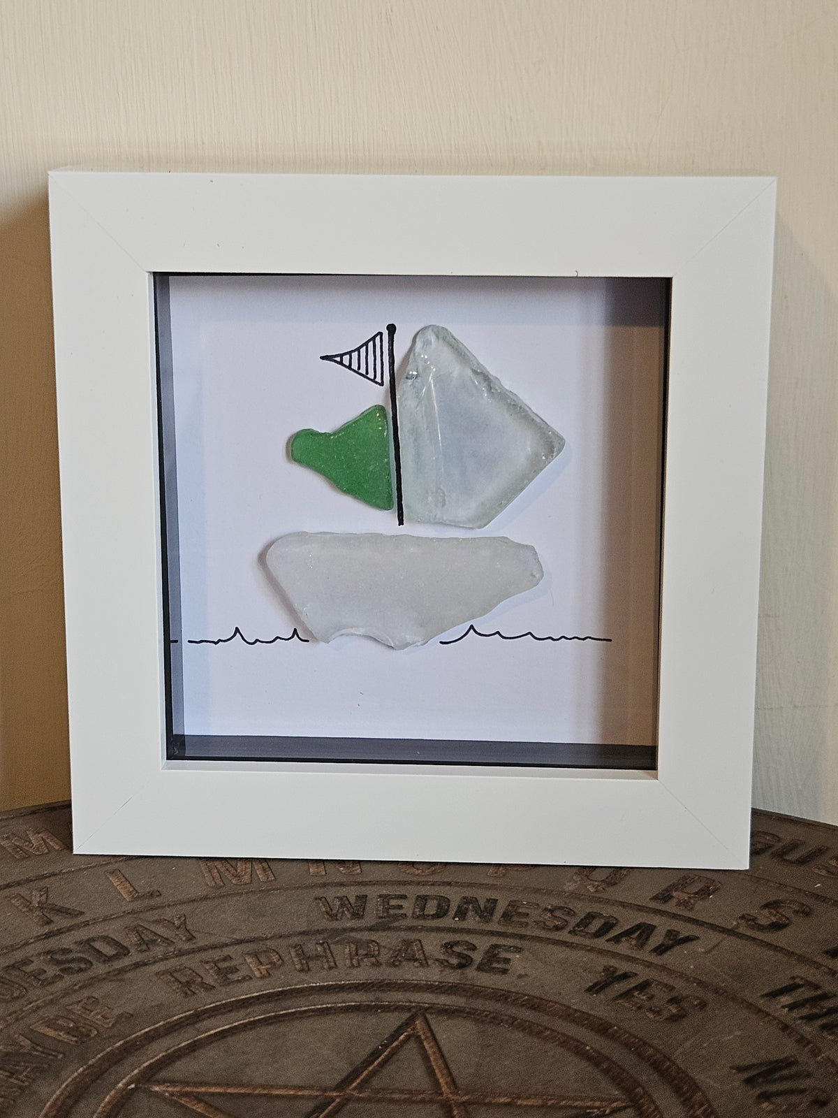 A square white shadow box framed piece of wall art featuring a simple nautical scene with two pieces of sea glass and a small black flag, displayed against a beige wall above a round wooden surface.