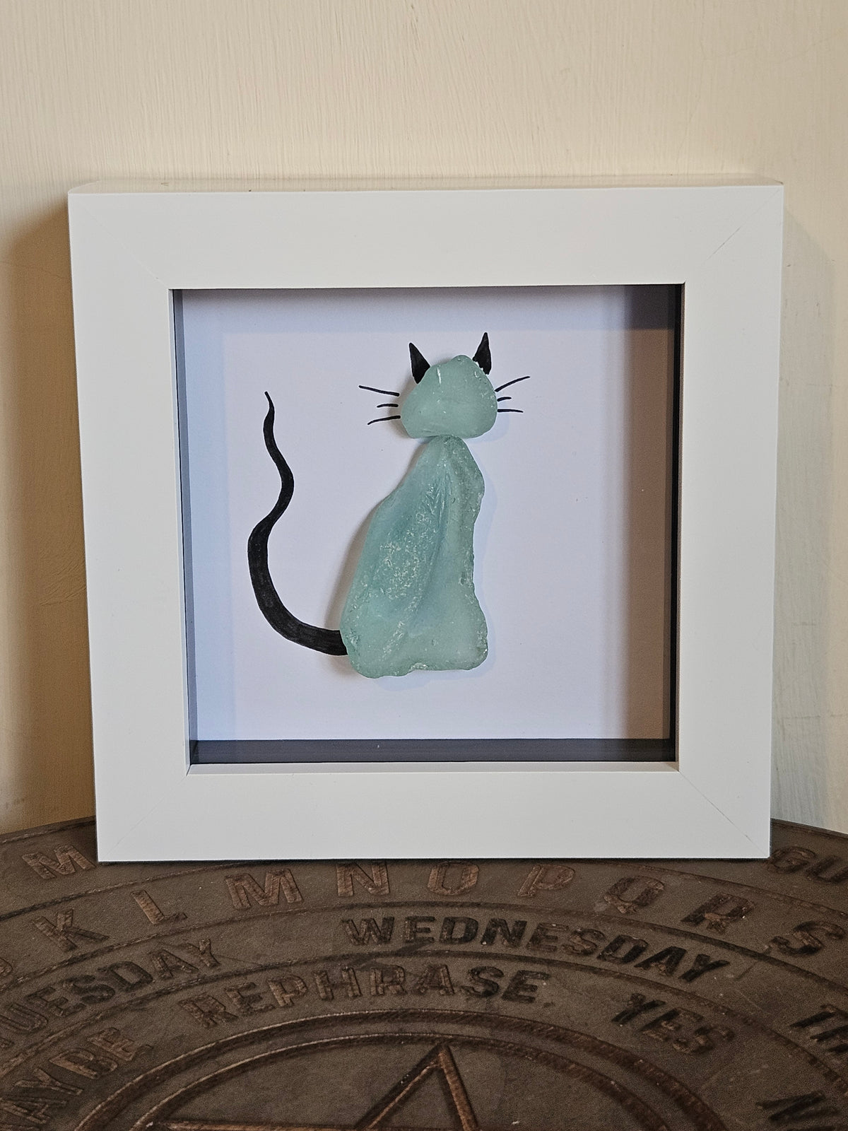 A framed shadow box art piece featuring a sculpted light blue-green cat with black ears, whiskers, and tail, displayed on a rustic wooden surface against a beige wall.