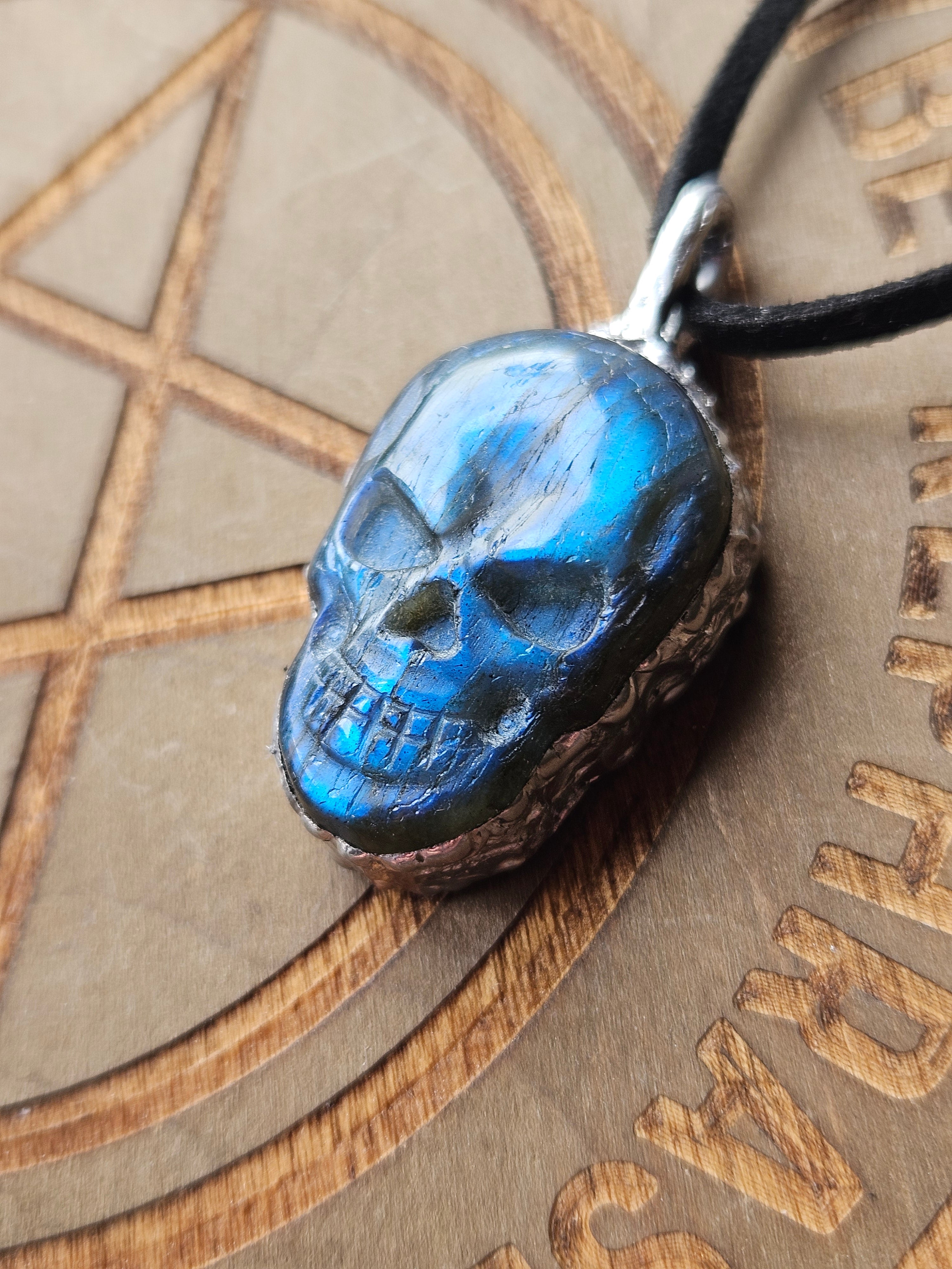 A close-up of a skull-shaped pendant necklace with a glossy blue finish and dark recessed details, hanging from a black cord against a brown engraved background.