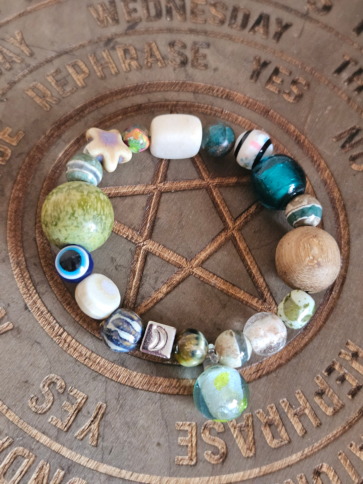 A handmade beaded boho bracelet arranged in a circle on a wooden surface featuring a carved pentagram and engraved words. The bracelet contains a mix of beads including a large green marbled sphere, a white rectangular bead, a star-shaped bead, and various blue, green, and patterned glass beads.
