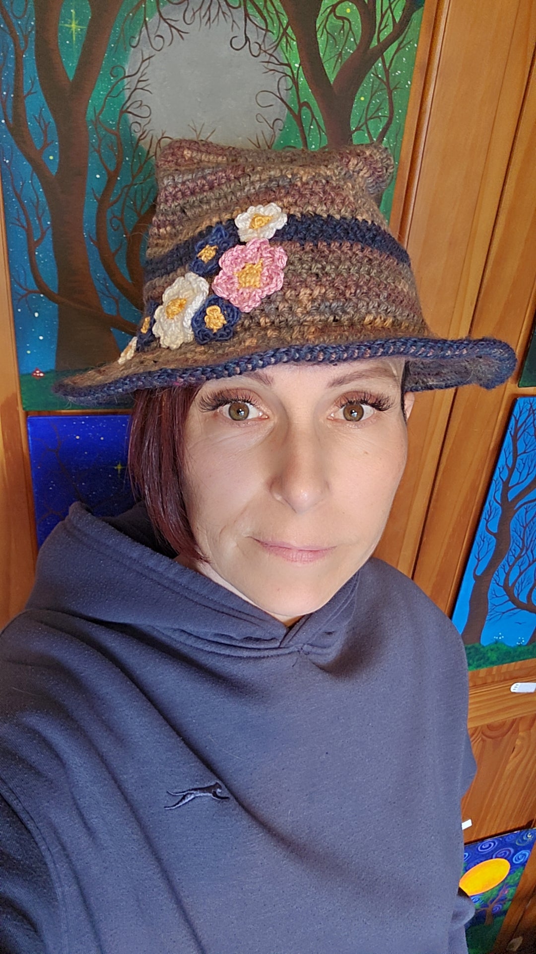 Person wearing a crocheted hat with floral decorations in front of a colorful wall.