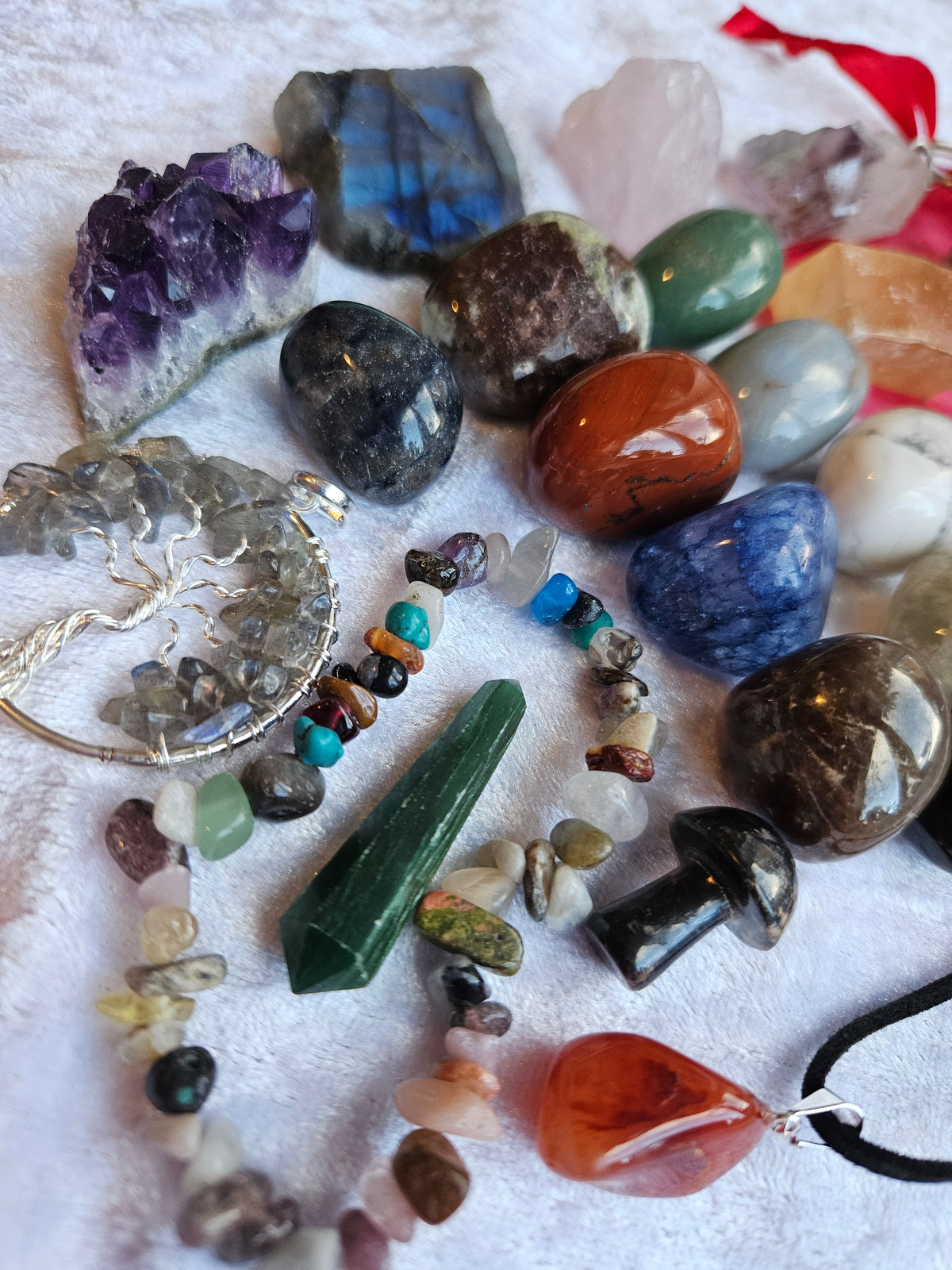 A collection of colorful raw gemstones, polished crystals, and beaded jewelry arranged on a white textured fabric background. The assortment includes a tree of life pendant, a teardrop-shaped amber pendant, a green cat's eye stone, a purple amethyst cluster, and various other polished stones and bead strands.