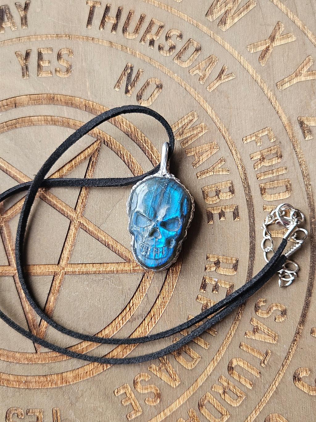 A skull-shaped pendant with a vibrant blue marbled inlay, set in a silver-toned metal frame and suspended from a black cord necklace, displayed on a wooden pentagram background.