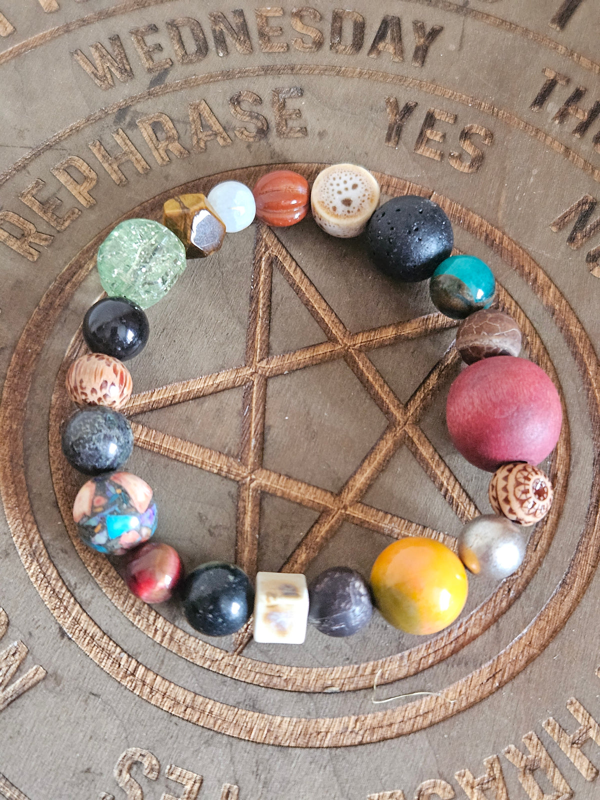 A beaded boho bracelet arranged in a circle on top of a carved wooden pentagram and calendar board. The beads are made from various natural stones and crystals in multiple colors including black, green, red, yellow, orange, and white.