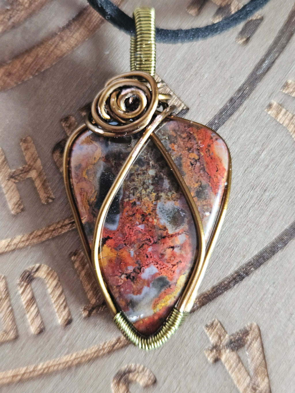 A close-up of a handmade wire-wrapped pendant featuring a polished natural stone with orange, red, brown, and black marbled patterns, set in an antique gold-tone metal frame with a spiral wire accent on the side.