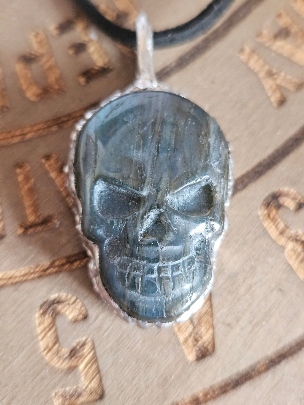 A skull-shaped pendant made of polished labradorite with a silver-toned bezel, resting on a wooden board engraved with symbols.
