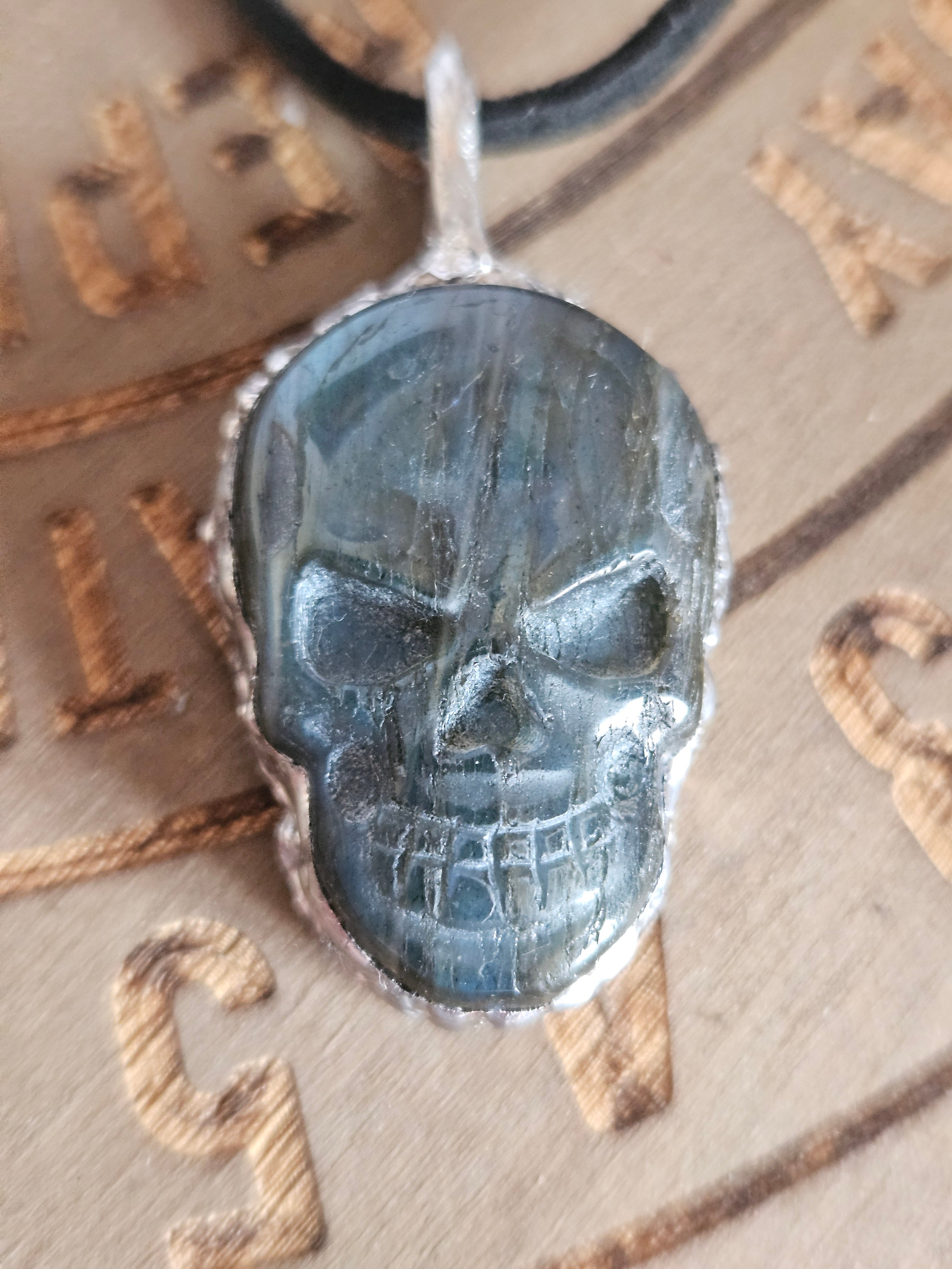 A skull-shaped pendant made of polished labradorite with a silver-toned bezel, resting on a wooden board engraved with symbols.