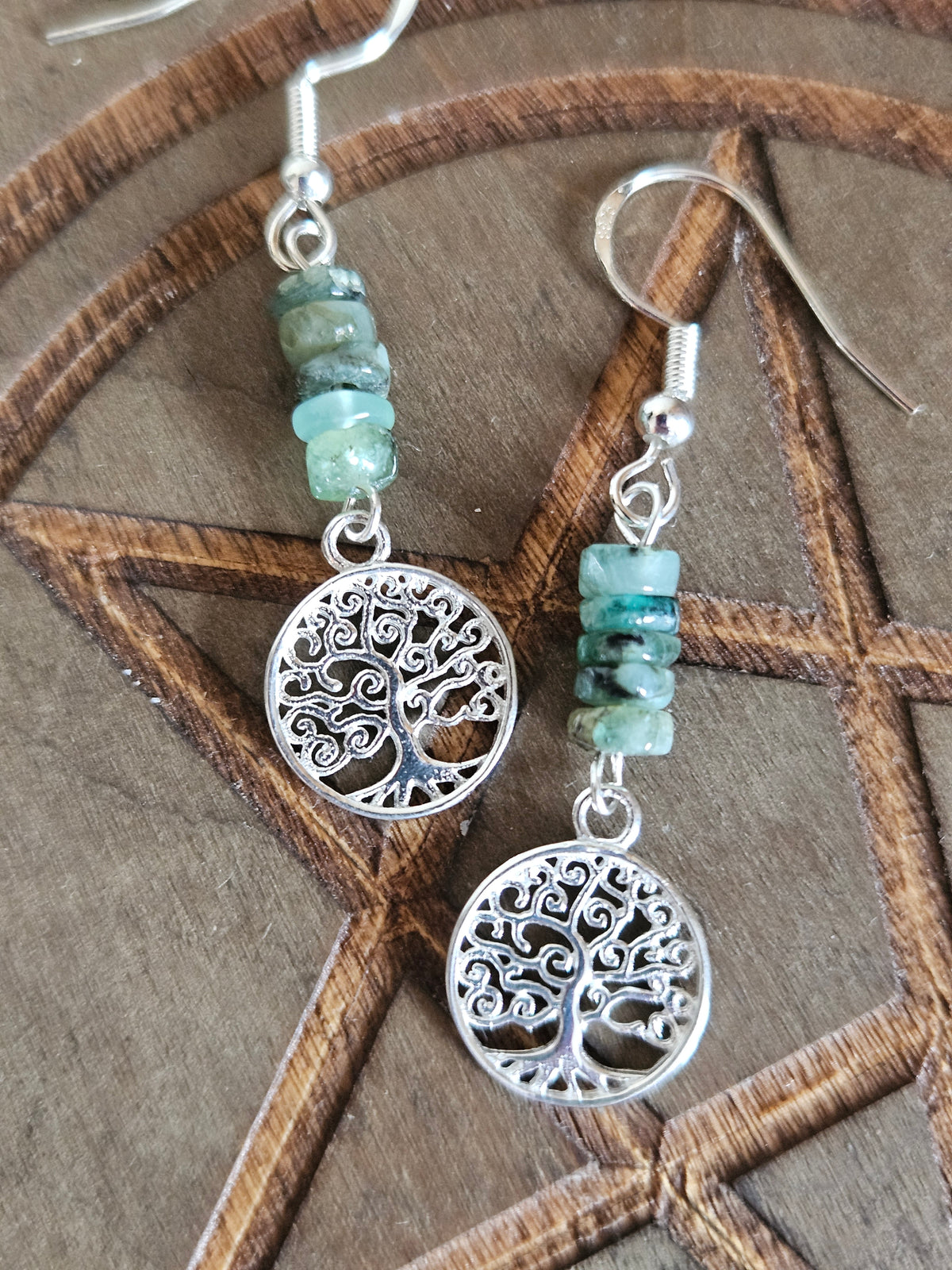 A pair of bohemian-style tree of life dangle earrings with sterling silver tree of life circles, green Emerald beads, and sterling silver fish hooks, displayed on a wooden pentagram background.