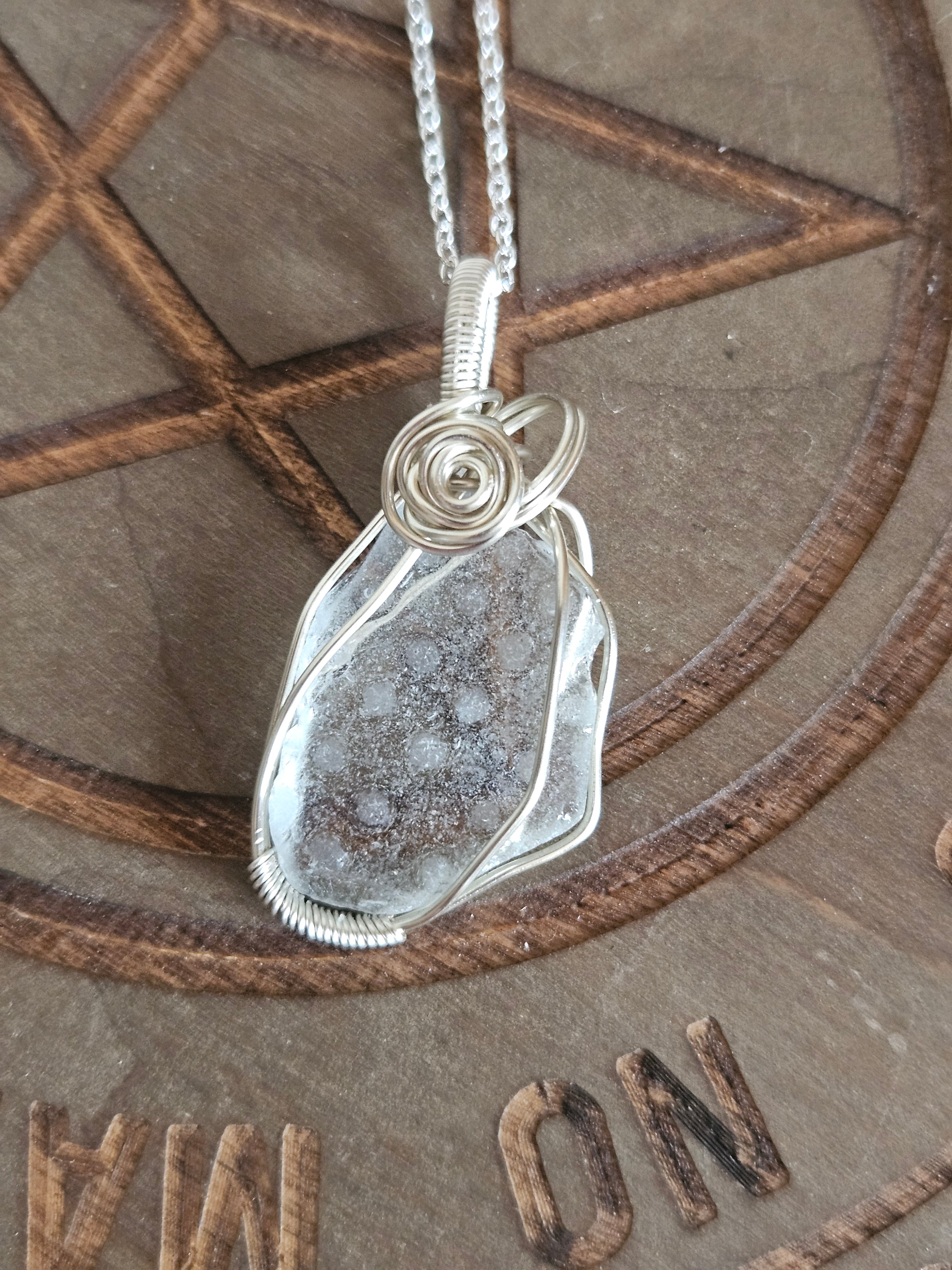A silver-wrapped pendant necklace featuring a frosted piece of polka dot patterned sea glass, displayed on a brown wooden pentagram background.