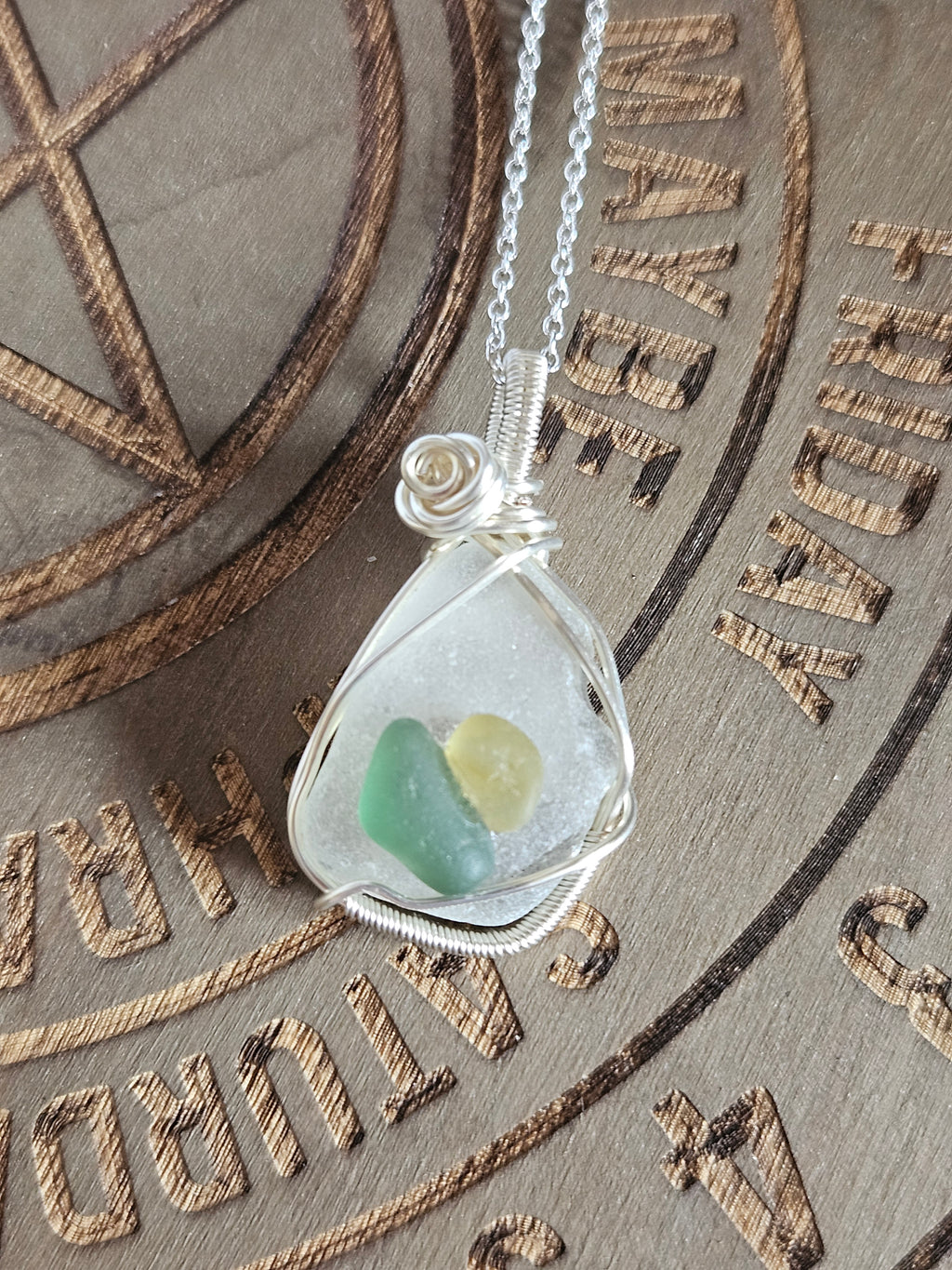A silver wire-wrapped necklace with a teardrop-shaped pendant containing small green and pale yellow sea glass, displayed on a brown engraved parchment background.