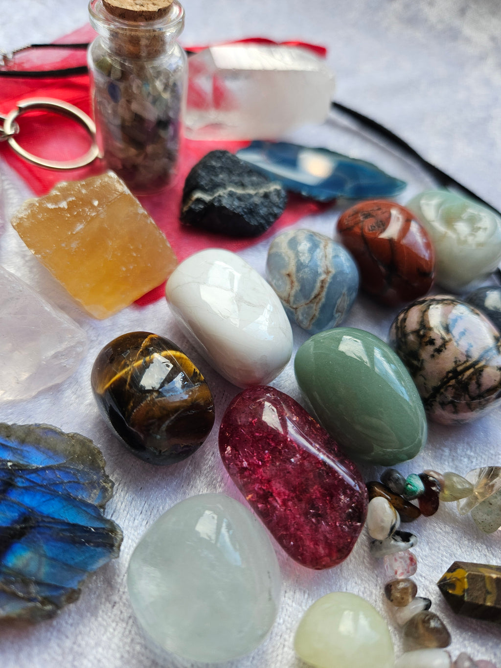 A collection of polished and raw gemstones and crystals arranged on a white textured fabric, including a small glass vial with a cork stopper, a red crystal, a blue labradorite, a tiger's eye, a white quartz, and other assorted stones.