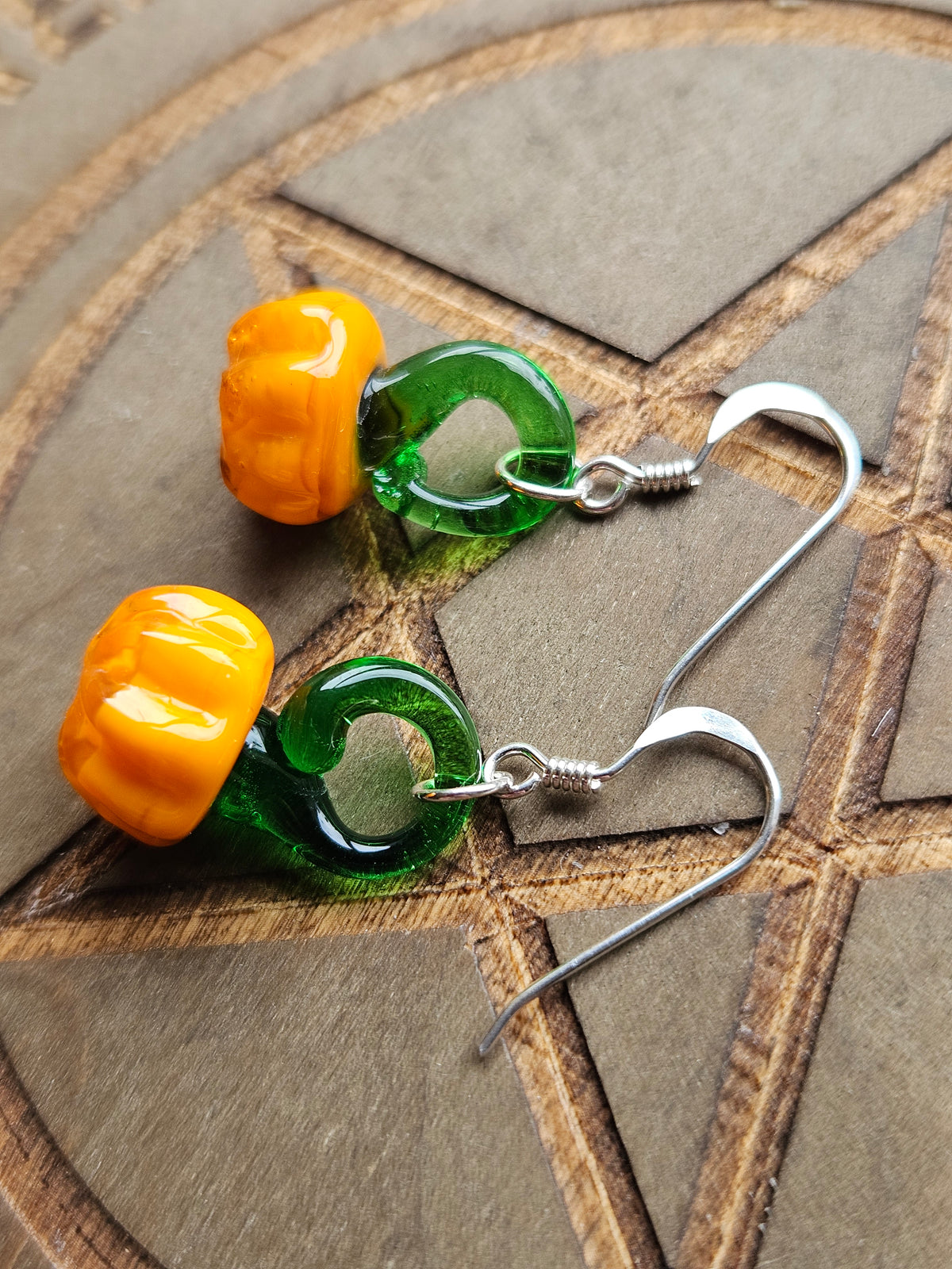 A pair of handmade dangle earrings featuring bright orange pumpkin-shaped beads connected to translucent green glass loops on silver-tone fishhook wires, displayed on a textured brown geometric surface.