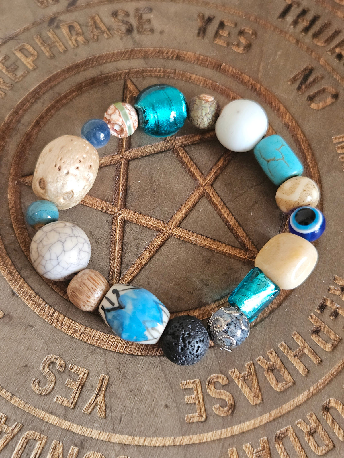 A beaded boho  bracelet with a variety of textured, polished, and patterned beads in turquoise, cream, white, and black, arranged in a circle on a wooden pentagram board.