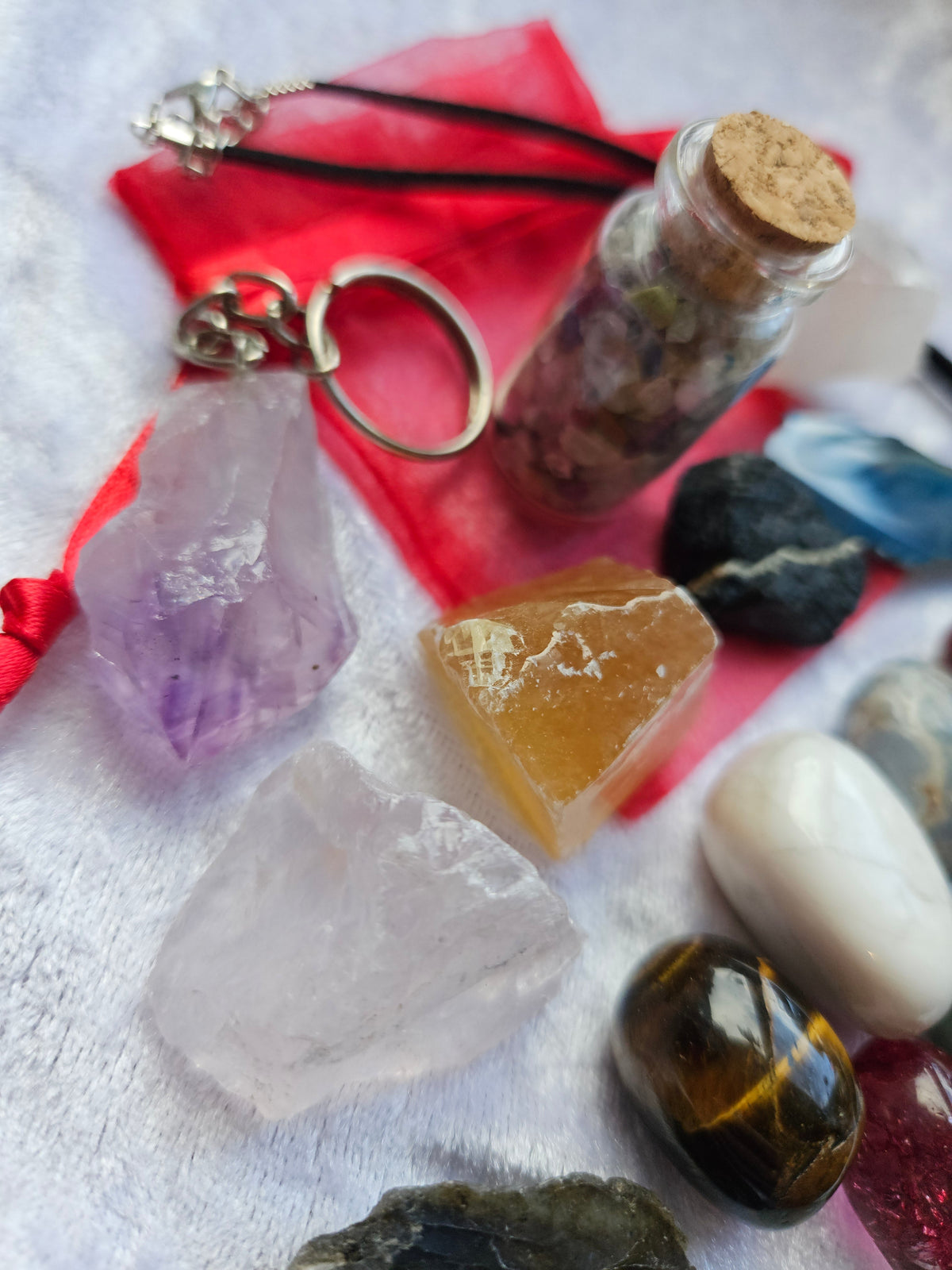 A collection of crystals and small accessories arranged on a white textured fabric. The set includes a small glass vial with a cork stopper, a red fabric pouch with a silver keychain, a black cord necklace with a silver charm, a translucent purple crystal, a pale pink crystal, a yellow-orange stone, and several polished and rough dark stones.