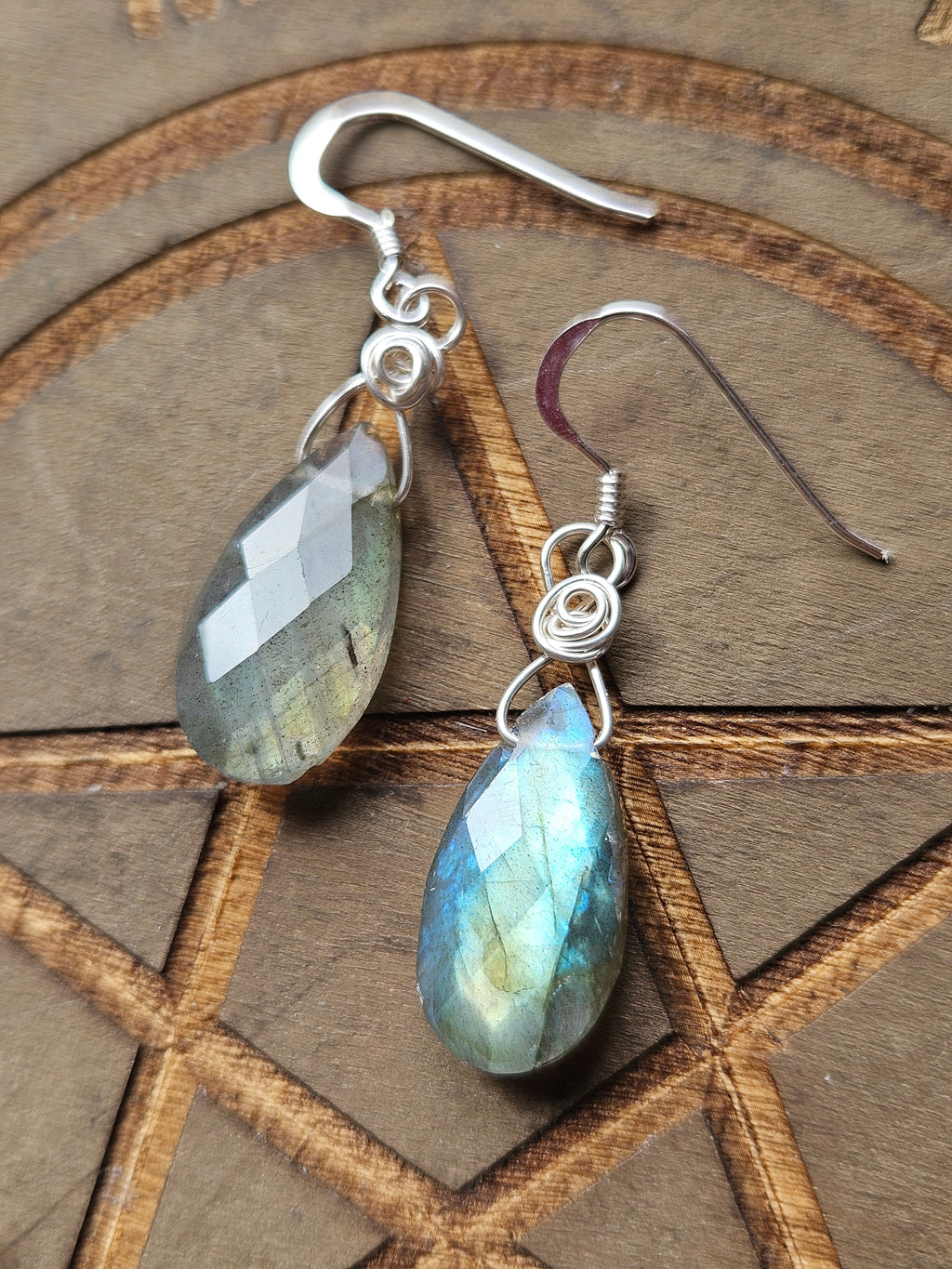 A pair of teardrop-shaped labradorite dangle earrings with silver wire-wrapping and silver fishhook ear wires, displayed on a wooden background with an engraved pentagram.