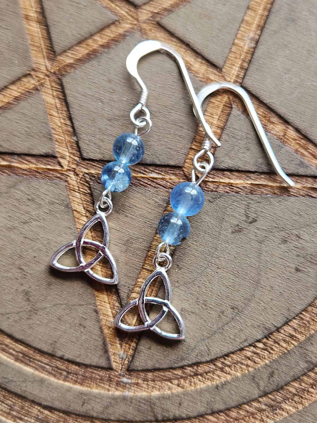 A pair of silver dangle earrings with blue Aquamarine beads and Sterling Silver Celtic knot charms, displayed on a wooden surface with a pentagram design.