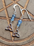 A pair of silver dangle earrings with blue Aquamarine beads and Sterling Silver Celtic knot charms, displayed on a wooden surface with a pentagram design.